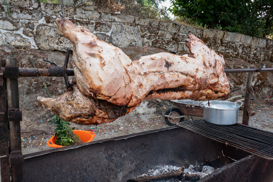 Porco no espeto a rodar sobre brasas num assador. Churrasco r&uacute;stico ao ar livre com uma panela ao lado