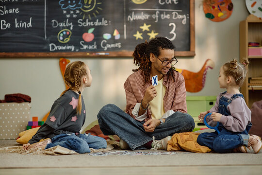 Caucasian girl and another Caucasian girl sitting on floor interacting with Black man teacher holding flashcard in kindergarten classroom, children leaning alphabet in educational environment