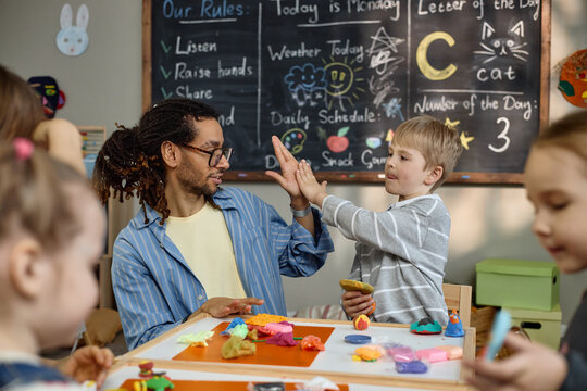 Caucasian boy giving high five to Black man teacher while sitting at table with colorful modeling clay, other children participating in creative activity in kindergarten classroom