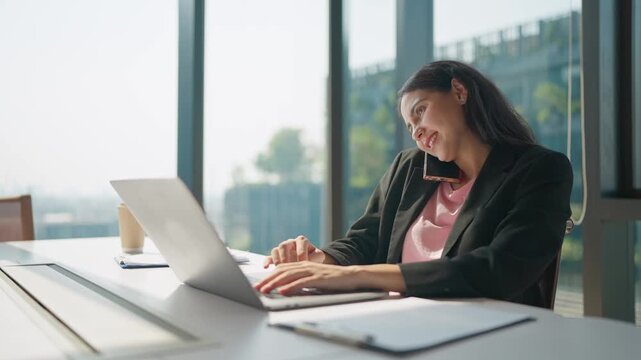 Businesswoman's Multitasking: A focused businesswoman juggles tasks, engaging in a phone call while skillfully working on her laptop in a modern office environment.