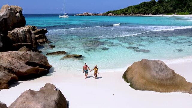 A couple strolls hand in hand along the pristine shores of Anse Cocos beach, enjoying the clear turquoise waters and scenic rock formations. A perfect tropical getaway in La Digue Seychelles awaits.