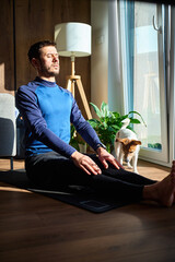 Man practicing breathing exercise while sitting on mat in sunny home interior with dog nearby....