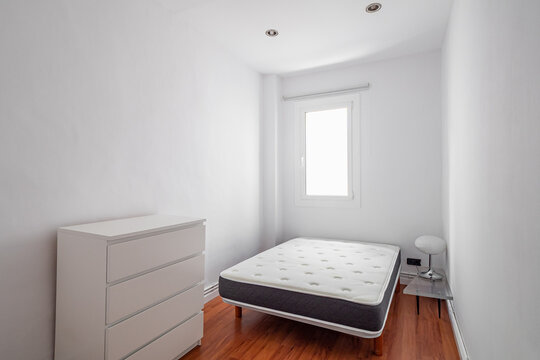 A minimalistic bedroom with a single bed and a white dresser. The room has white walls and wooden flooring, with a window allowing natural light.