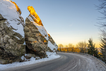 Fototapeta premium A winding road wraps around a large rock covered in snow. The sun sets in the background, casting light on the rock. Trees stand bare around the road during winter.