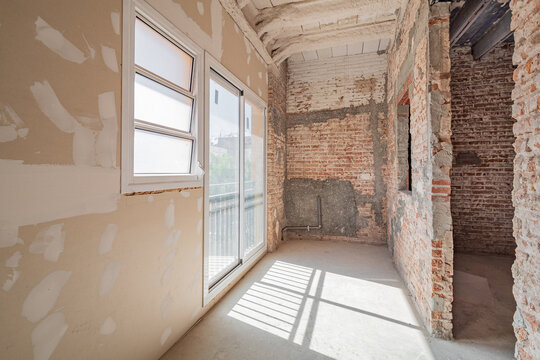 An unfinished room undergoing renovation. Exposed brick walls and drywall are visible. Natural light enters through a large window, illuminating the space.