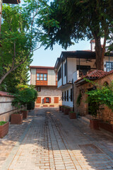Fototapeta premium Antalya, Turkey. Quiet residential street in Kaleici Old Town with cobblestone pavement, white two-story buildings, red tiled roofs, red door, and trees casting shadows on sunny morning.
