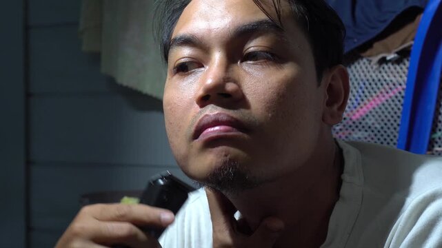 Side view and close-up of a young Asian man using an electric hair trimmer to shave his beard and neck. Professional male grooming routine, skincare, and daily hygiene at home. Highlighting facial hai