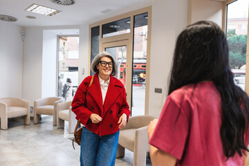 Woman in red jacket arriving at a modern dental clinic, being greeted by a receptionist wearing scrubs. Healthcare appointment © Koldo_Studio