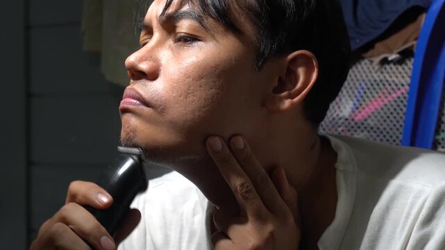 Close up of a Southeast Asian man trimming his beard and mustache with an electric hair clipper. Concept of authentic male grooming, skincare challenges with acne scars, and daily hygiene routine in a