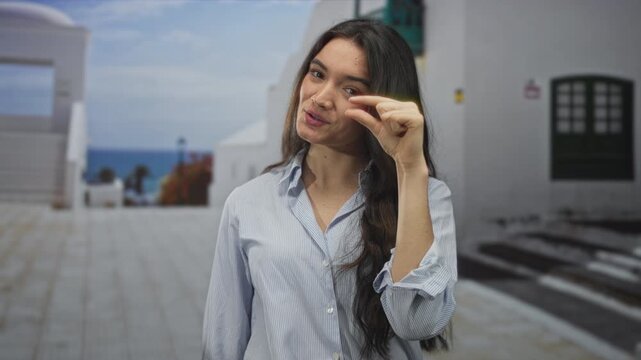 Young hispanic woman pinches fingers to show small size in front of a street building under bright sunlight; precision.