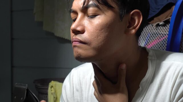 Close-up of focused Asian man with acne scars using an electric clipper to shave his beard and neck hair. Detailed 4K video showing men's grooming routine, personal hygiene, and male skincare in a hom