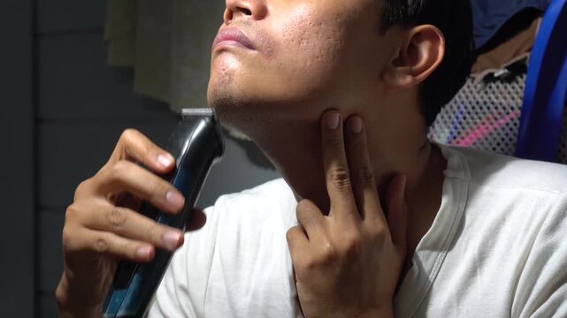 Close up of a young Asian man shaving his chin and neck with an electric trimmer clipper, checking his skin texture afterwards.