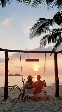 A couple enjoys a serene moment on a swing overlooking the vibrant sunset in Koh Chang, Thailand. The beach setting is tranquil, offering a perfect escape for lovers and travelers seeking relaxation.