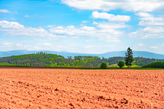 The scene shows a large area of agricultural land featuring red clay soil in the vicinity of Nova Paka, Czechia. Trees and hills are visible in the background under a bright sky.