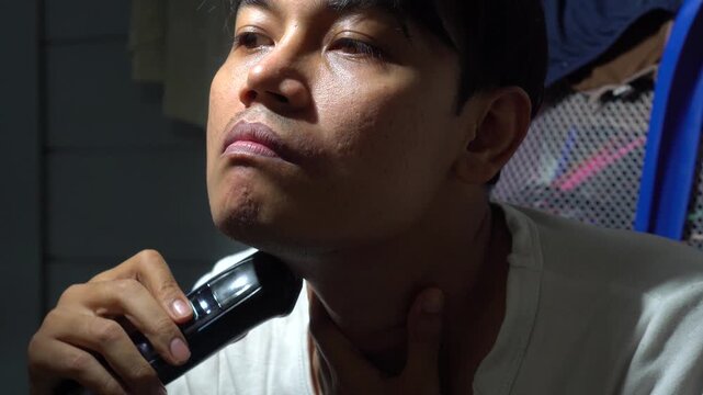 Asian man shaving his beard and facial hair using an electric trimmer at home. Concept for men grooming, skin care routine, hygiene, personal care, acne skin texture, and daily lifestyle.