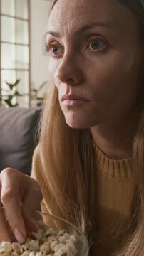 Vertical close-up of face of startled young Caucasian woman watching slasher movie or thriller on TV, with vivid emotions and wide eyes, shocked by violent or explicit content, eating popcorn