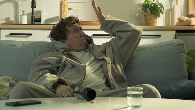 Young man waking up on couch in modern apartment and immediately taking a pill with water. Daily habit, need for relief and tired lifestyle at home, ambiguous situation between routine and dependence