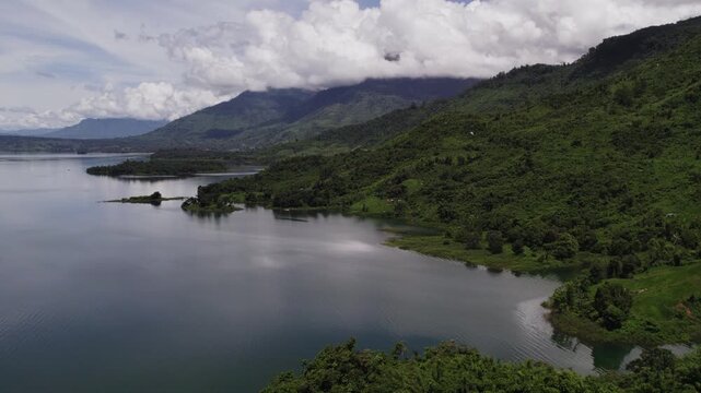 Flying high altitude forward moving view of coastline of tropical lake with lish tropical vegetation covered mountains and cloudy blue sky in the background.