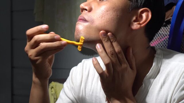 Asian man shaving facial hair with a yellow manual razor blade at home. Concept for men grooming routine, acne skin texture care, personal hygiene, daily shaving, beard trimming, morning routine, and 