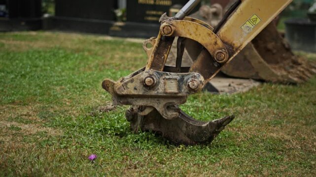 A frustrating and tedious moment as a small excavator repeatedly attempts to connect a hook attachment to its arm while operating on grass in a cemetery.