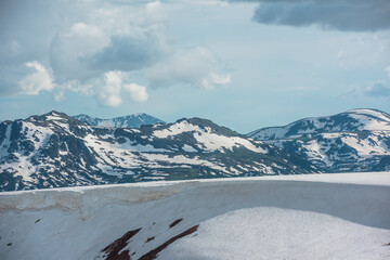 Scenic alpine landscape with sunlit snowfield with snow cornice above abyss against large snow-capped rock mountain top. Sunlight and shadows on snowy field near rocky precipice edge in high mountains © Daniil