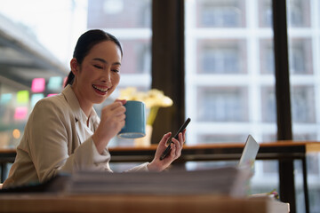 Fototapeta premium Young Asian businesswoman smiling and joyfully interacting with a mobile phone while holding a mug, taking a break from work at her desk in a modern office setup