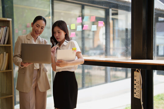 Two Asian businesswomen are collaborating, discussing, and planning new strategies using a laptop during a meeting in a modern office with sticky notes on a glass wall