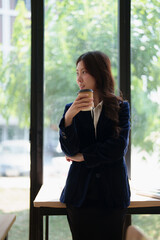 Fototapeta premium Young Asian businesswoman in formalwear stands by a large window with a disposable coffee cup, gazing thoughtfully as she plans career growth and future success in a modern office