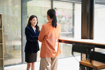 Fototapeta premium Two Asian businesswomen are collaborating and networking during a casual meeting in a modern office, standing and discussing a project while one is smiling