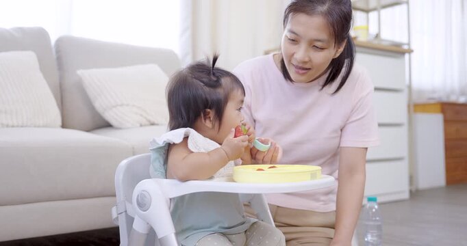 Asian mother interacts with toddler girl during breakfast, child holds spoon fruit, smiling softly, bright morning light across cozy room, nurturing love, joyful self feeding, early development moment