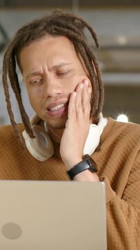 Vertical video: Reacting to laptop, dreadlocked man in sweater rubbing jaw at desk, with headphones