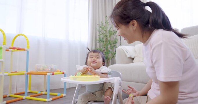 Asian mother kneeling beside toddler girl sitting on feeding chair, smiling and talking during mealtime, bright natural light fills cozy room, nurturing connection, family bonding, early learning