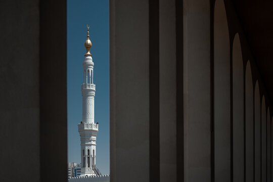 Minaret of a mosque in downtown Abu Dhabi, United Arab Emirates, framed between shaded colonnades with the white tower rising against a clear blue sky.