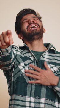 Amused bearded Indian man pointing finger to camera, laughing out loud, taunting making fun of ridiculous appearance, funny joke anecdote. Handsome Hindu boy isolated alone on beige studio background