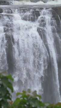 slow motion video of water fall crashes over a steep cliff. Rushing torrents form a frothy, white curtain. Green trees frame the distant, misty horizon. Powerful nature dominates the scene with raw