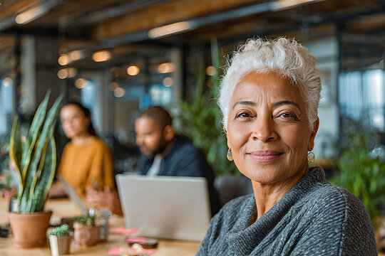 Confident mature professional woman smiling in a modern coworking office - senior leader portrait with colleagues and greenery in bright workspace