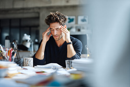 Young man overwhelmed at a cluttered desk, holding his temples &mdash; stressed professional struggling with paperwork, deadlines and creative burnout in office