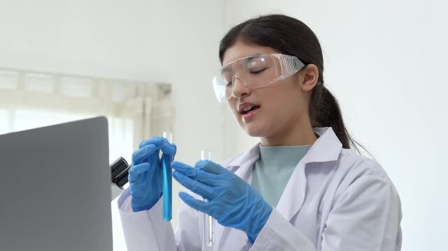 Young Asian women scientist working with test tube in laboratory. Scientist in a lab coat and safety glasses handles a test tube at a desk in a laboratory, make a verbal note on laptop experimentation