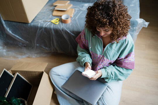 Woman sitting on floor at home using smartphone while unpacking