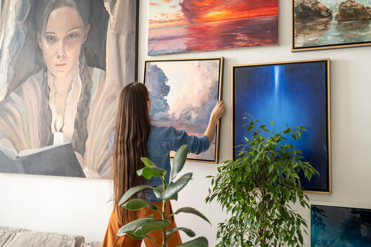 Talented woman painter hanging new finished artwork canvas on wall at home loft gallery, showcasing skills and love for art. Successful female artist arranging own exhibition in studio living room.