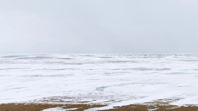 Minimal winter beach facing a frozen sea under a pale gray sky. Snow patches on sand and a flat horizon create strong copy space for cold, remote mood.