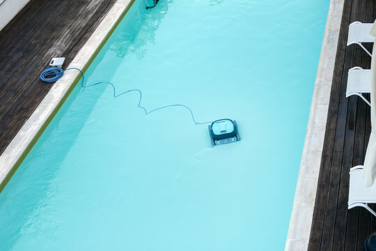 Robotic pool cleaner in outdoor swimming pool with wooden deck and lounge chairs, automatic cleaning technology concept with clear blue water and copy space