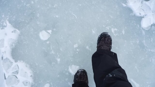 POV boots stepping across smooth pale blue ice with scattered snow chunks, showing risky winter walking on a frozen lake or sea. Minimal frame with copy space for safety warning.