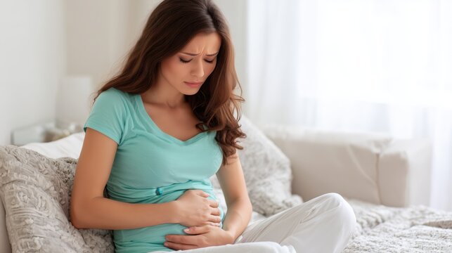A pregnant woman sits on a sofa, looking down at her belly with an expression of discomfort or pain.