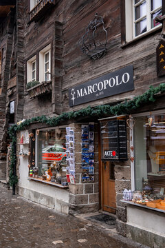 Rustic wooden MARCOPOLO shop, established in 1971, stands in Zermatt, Switzerland, with postcards, souvenirs, and treats by the entrance under overcast winter light.