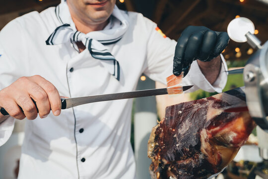 Men's hands with a sharp knife very thinly and skillfully cut Jamon meat on the street during the day.