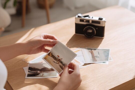 Nostalgia concept with woman looking at old printed photographs on wooden table near window. Retro camera and warm natural light create cozy mood.