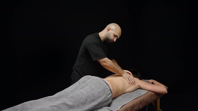 Professional massage therapist giving a soothing back massage to a client lying prone under a gray towel, applying hand techniques for muscle relief in a spa studio setting