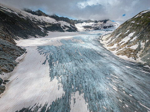 Aerial view of the Rhone Glacier's icy expanse, a river of blue and white snaking through the rugged, snow-dusted mountains, Obergoms, Valais, Switzerland.