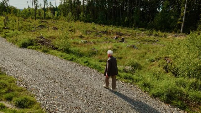 Elderly woman walking on forest path enjoying peaceful nature. Senior woman strolling along gravel road feeling calm. Retiree walking through woodland trail appreciating green landscape